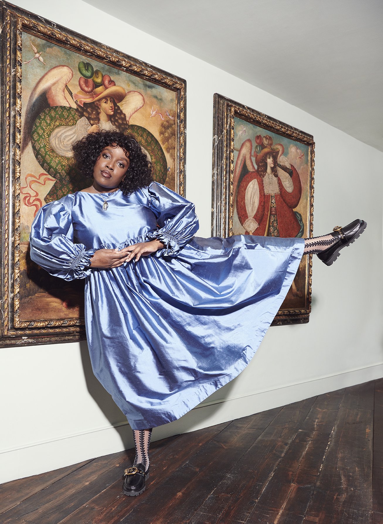Woman posing in front of two angel paintings in a gallery. She wears a light blue dress, patterned stockings, and black loafers.
