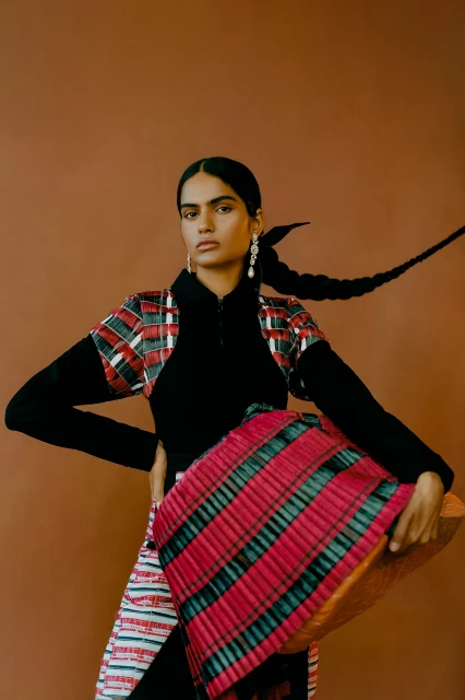 Portrait of a woman wearing a black turtleneck and a red plaid top holding a woven red and black patterned basket in front of a brown background.