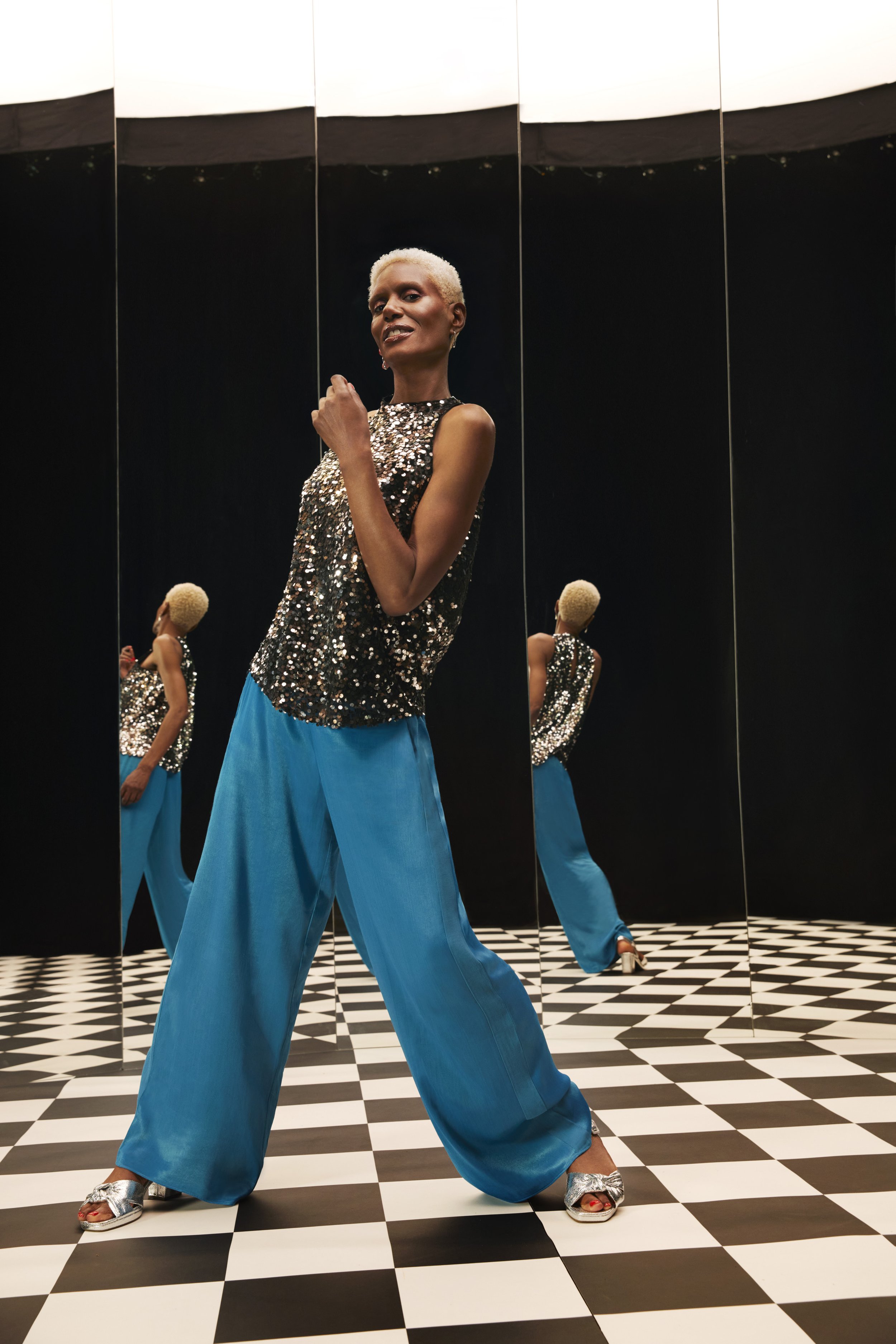 Woman in sequin top and wide-leg pants poses in a mirrored room with a black and white checkered floor, fashion photography.