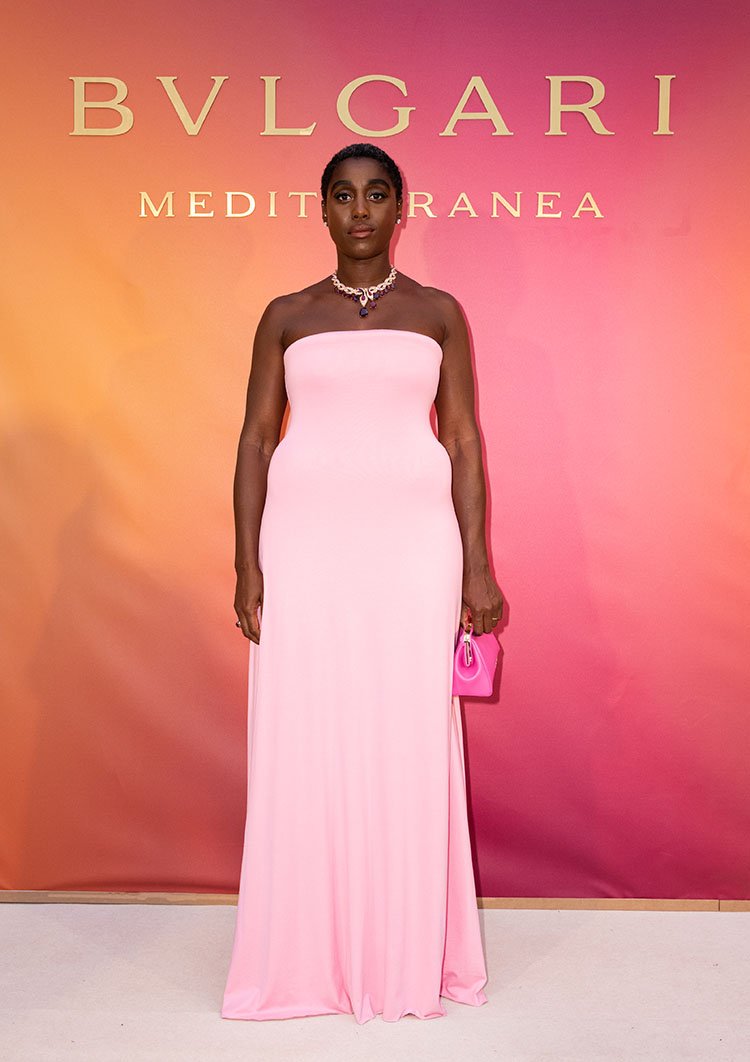 A woman with dark hair in a pink dress stands in front of a BVLGARI backdrop, holding a pink purse, at a fashion event.