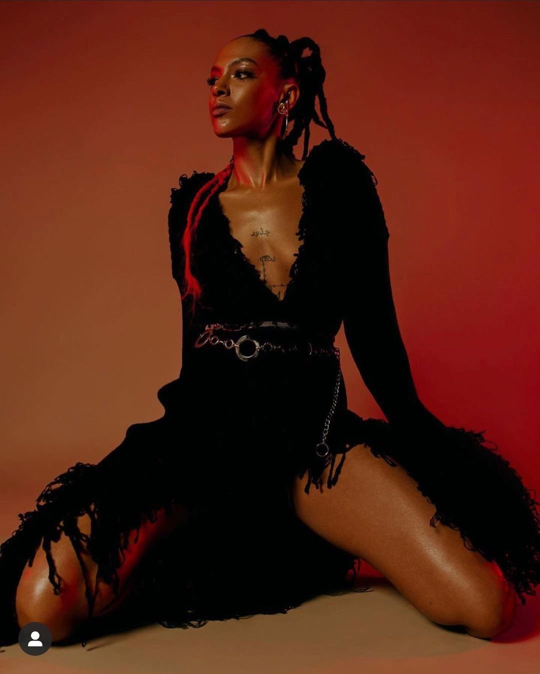 An adult woman is kneeling in a photo studio wearing a black dress with fringe and a chain belt, under soft lighting against a brown backdrop.