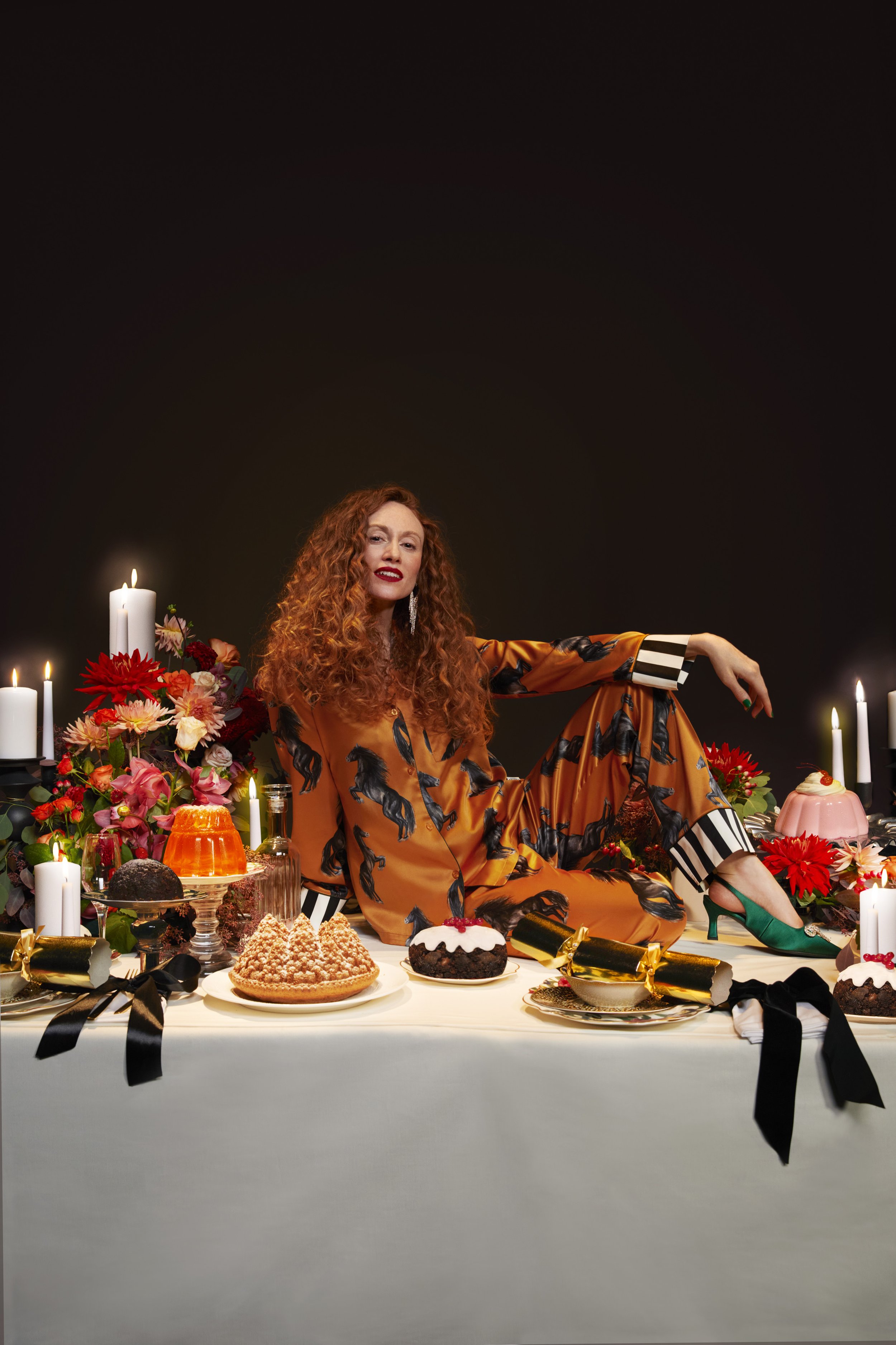 A woman with red curly hair, wearing a patterned pajama set, sits at a table filled with cakes, candles, and flowers in a dimly lit room.