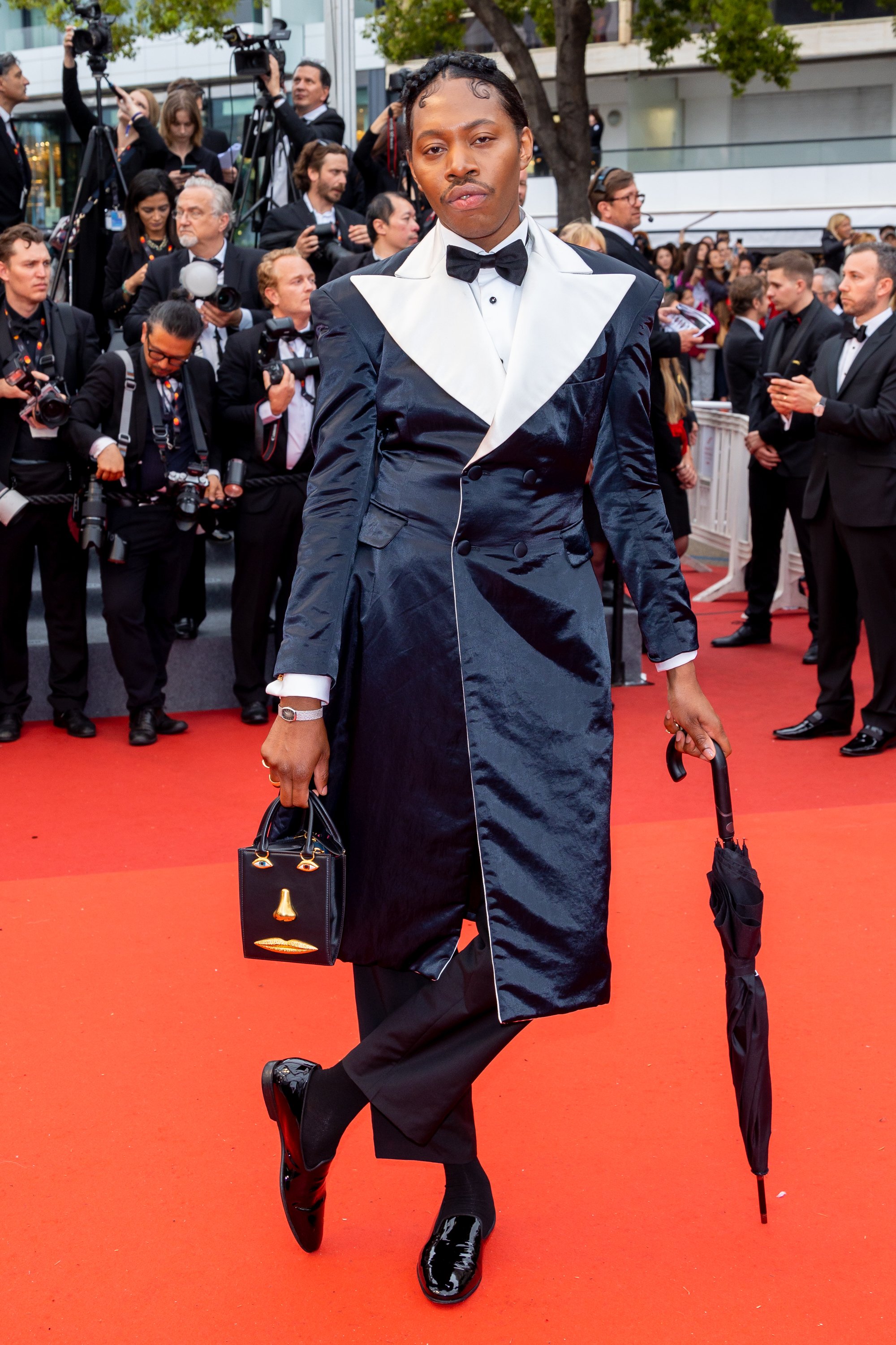 An individual poses on a red carpet, wearing a black and white tuxedo-style coat dress, holding a small purse and an umbrella.