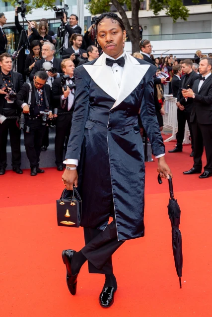 An individual poses on a red carpet, wearing a black and white tuxedo-style coat dress, holding a small purse and an umbrella.