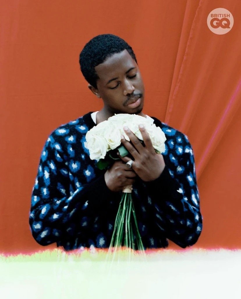 Portrait of a black male in patterned sweater holding white roses against a coral background. He gazes downward in a serene pose.
