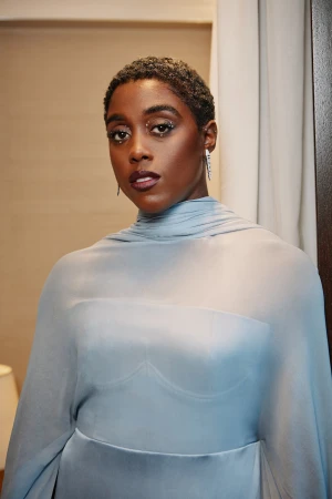Portrait of Lashana Lynch with short dark hair wearing a light blue sheer high neck dress with makeup by Min Sandhu