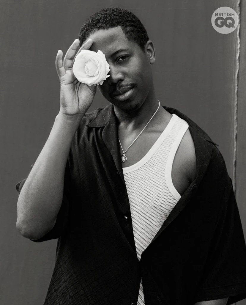 A stylish portrait of a young adult male in a black shirt, holding a white rose to his face, set against a dark background.