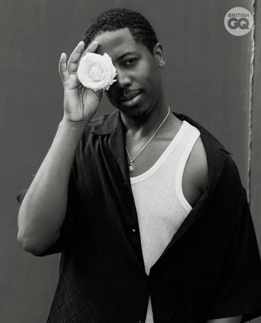 A stylish portrait of a young adult male in a black shirt, holding a white rose to his face, set against a dark background.