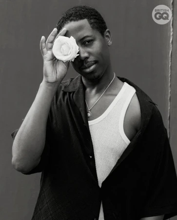 A stylish portrait of a young adult male in a black shirt, holding a white rose to his face, set against a dark background.