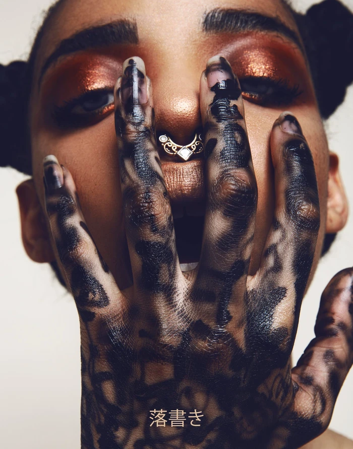 Close-up portrait of a woman holding her hand with intricate black ink markings across fingers in front of her face with copper eyeshadow.
