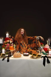 Red-haired woman in orange patterned silk pajama set sitting on a table with candles, cakes, and festive crackers.