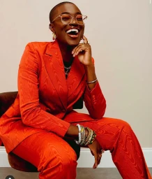 Black woman with a shaved head wearing a vibrant red patterned pantsuit and clear glasses laughing heartily.