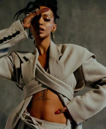 Woman in structured beige blazer posing with hand on forehead against a dark textured concrete wall background
