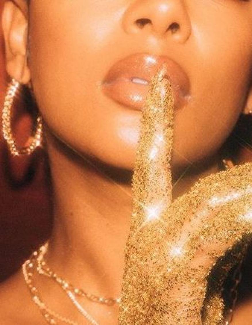 Close-up of a model with gold glitter-covered hand over the mouth, shiny lips, and gold jewelry against an orange background.
