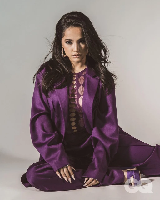Portrait of a woman with long dark hair wearing a purple suit, sitting cross-legged indoors against a light-colored backdrop, studio lighting.