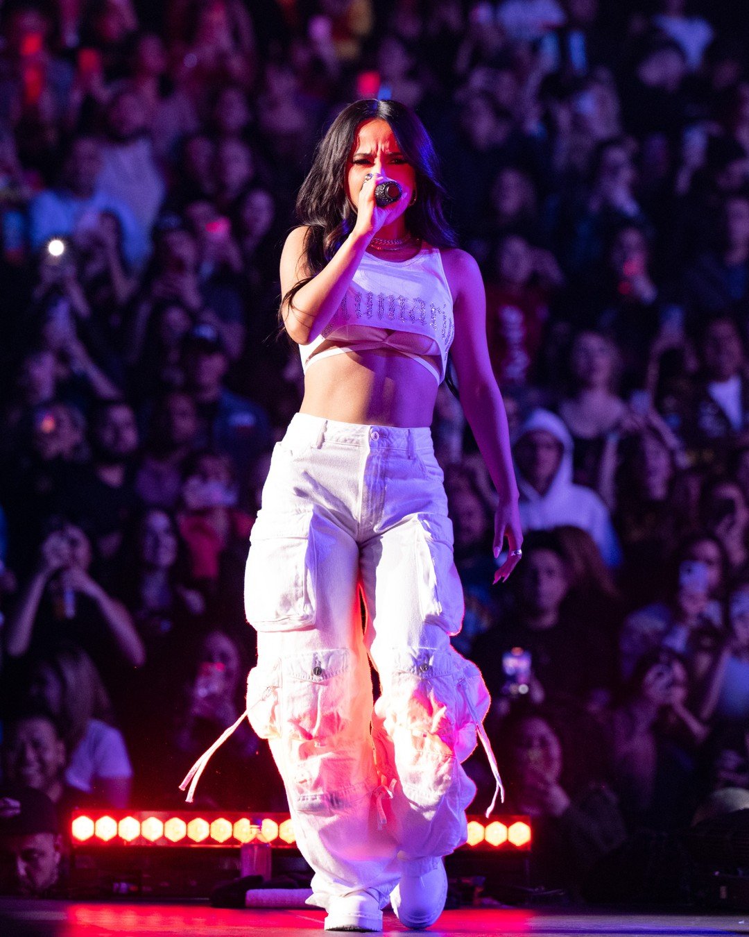 Singer performing on stage in a white crop top and baggy pants in front of a large crowd with stage lighting at night.