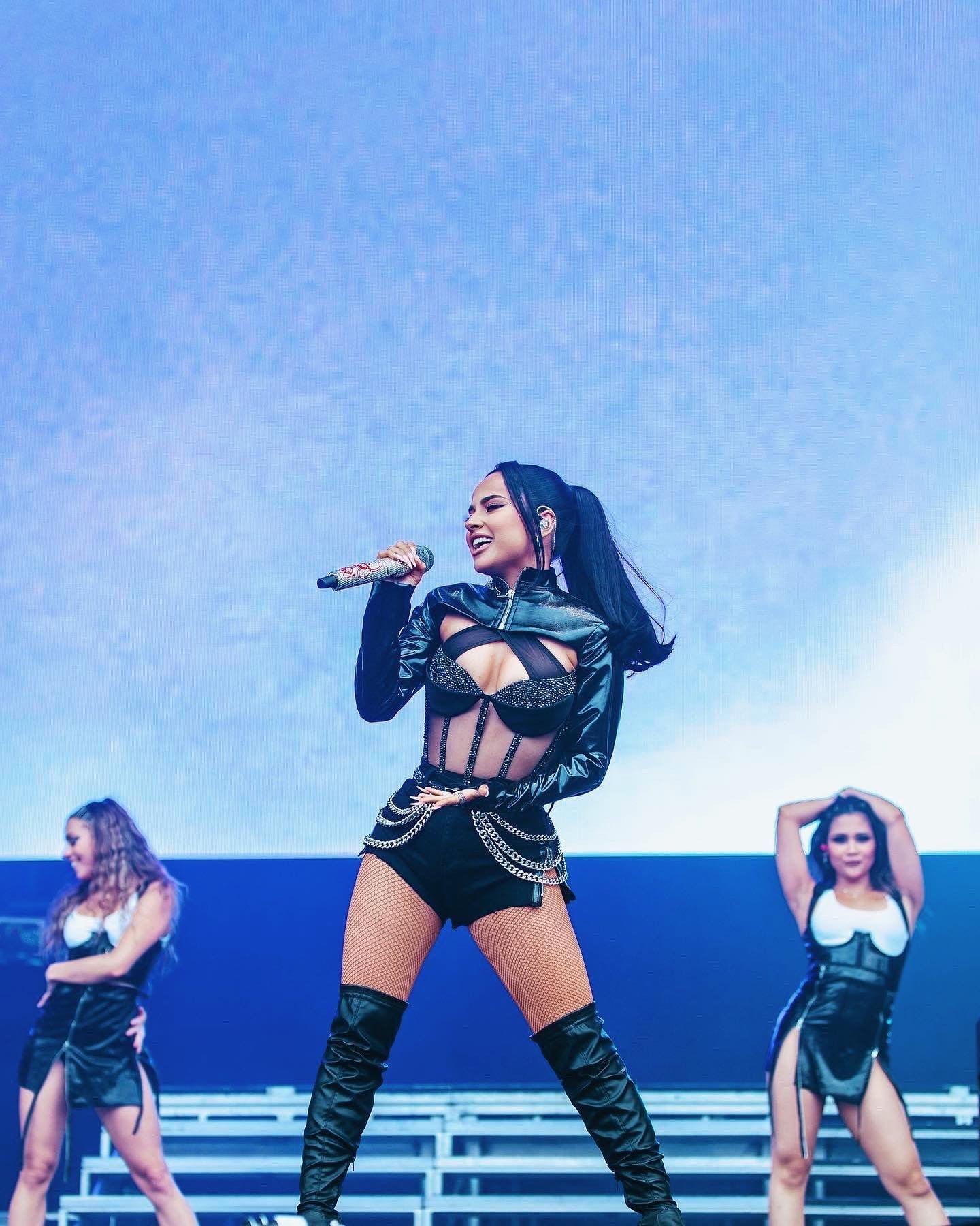 Female singer performing on stage at a concert in black outfit with backup dancers in a music event, singing into a microphone.