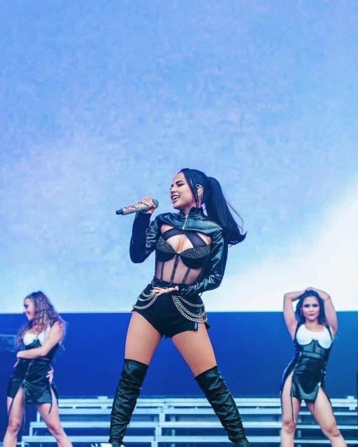 Female singer performing on stage at a concert in black outfit with backup dancers in a music event, singing into a microphone.