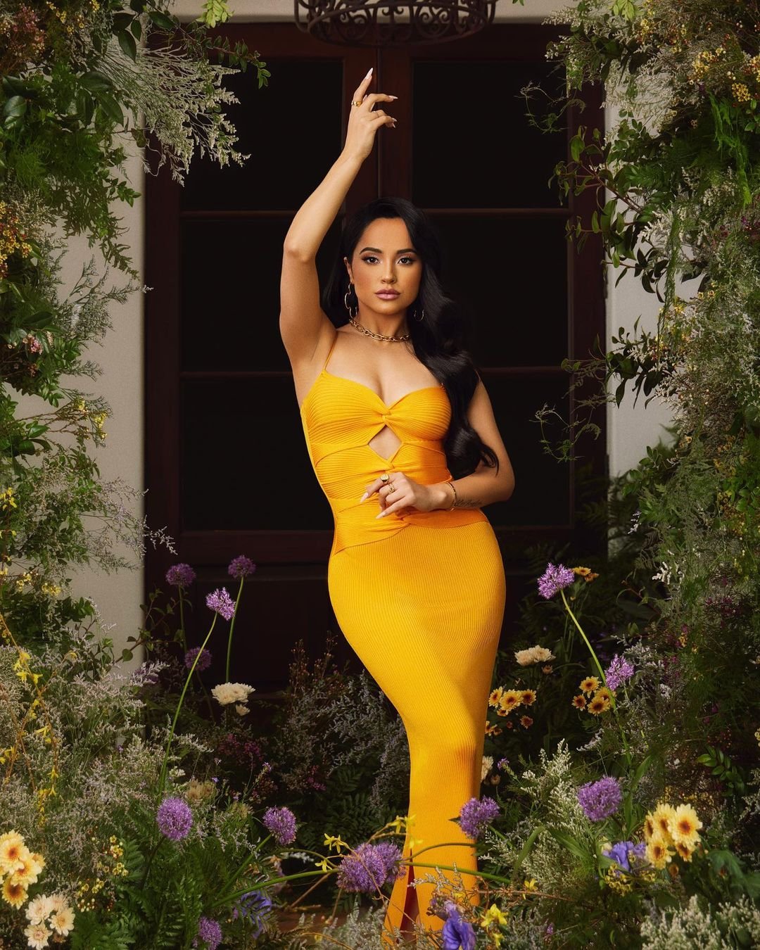 Adult female in a yellow gown poses in front of a door surrounded by flowering plants, wearing jewelry and with her arm raised.