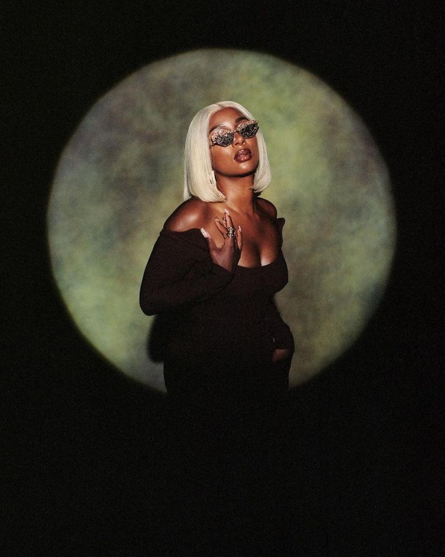 Glamorous Black woman with blonde bob and ornamental sunglasses posing in front of a spotlight casting a halo effect in a dark studio.