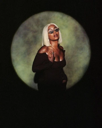 Glamorous Black woman with blonde bob and ornamental sunglasses posing in front of a spotlight casting a halo effect in a dark studio.