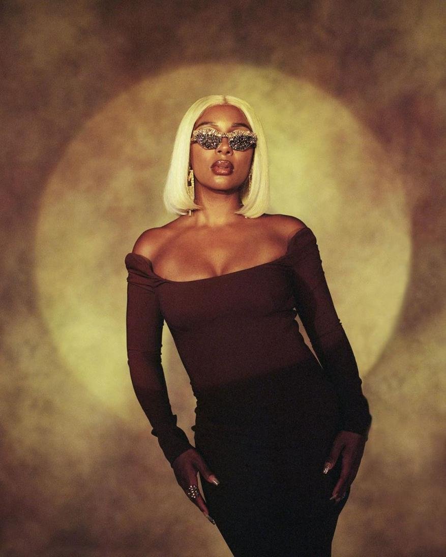 Portrait of an adult Black woman wearing an auburn dress, sparkling sunglasses, and blond wig, posing in a photo studio with warm lighting.
