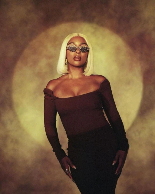 Portrait of an adult Black woman wearing an auburn dress, sparkling sunglasses, and blond wig, posing in a photo studio with warm lighting.