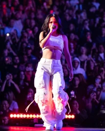 A female singer wearing a white crop top and cargo pants performs on stage with a blurred crowd in the background.