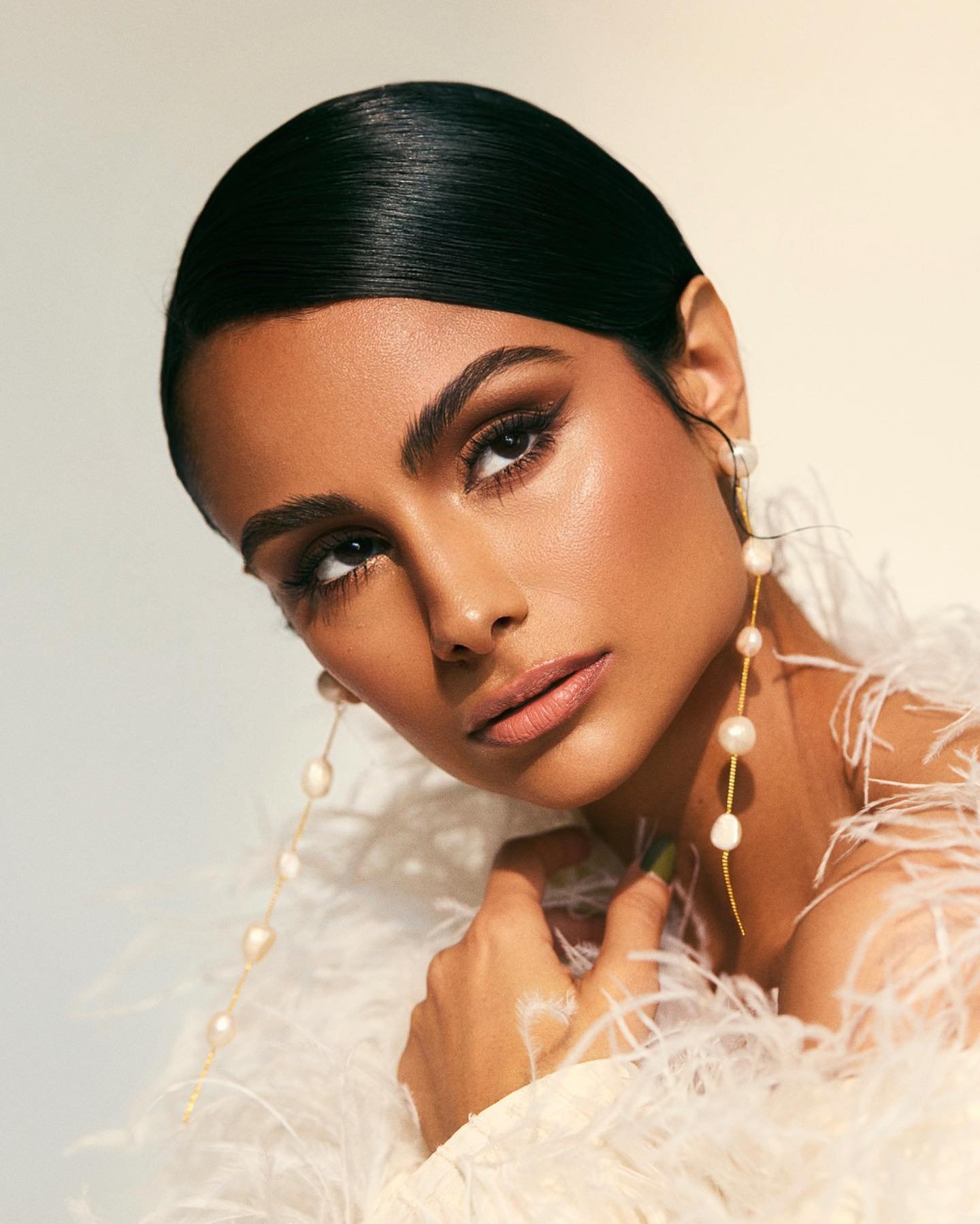Close-up of a woman with smooth, dark hair and glamorous makeup, wearing dangling pearl earrings and a feathered boa.