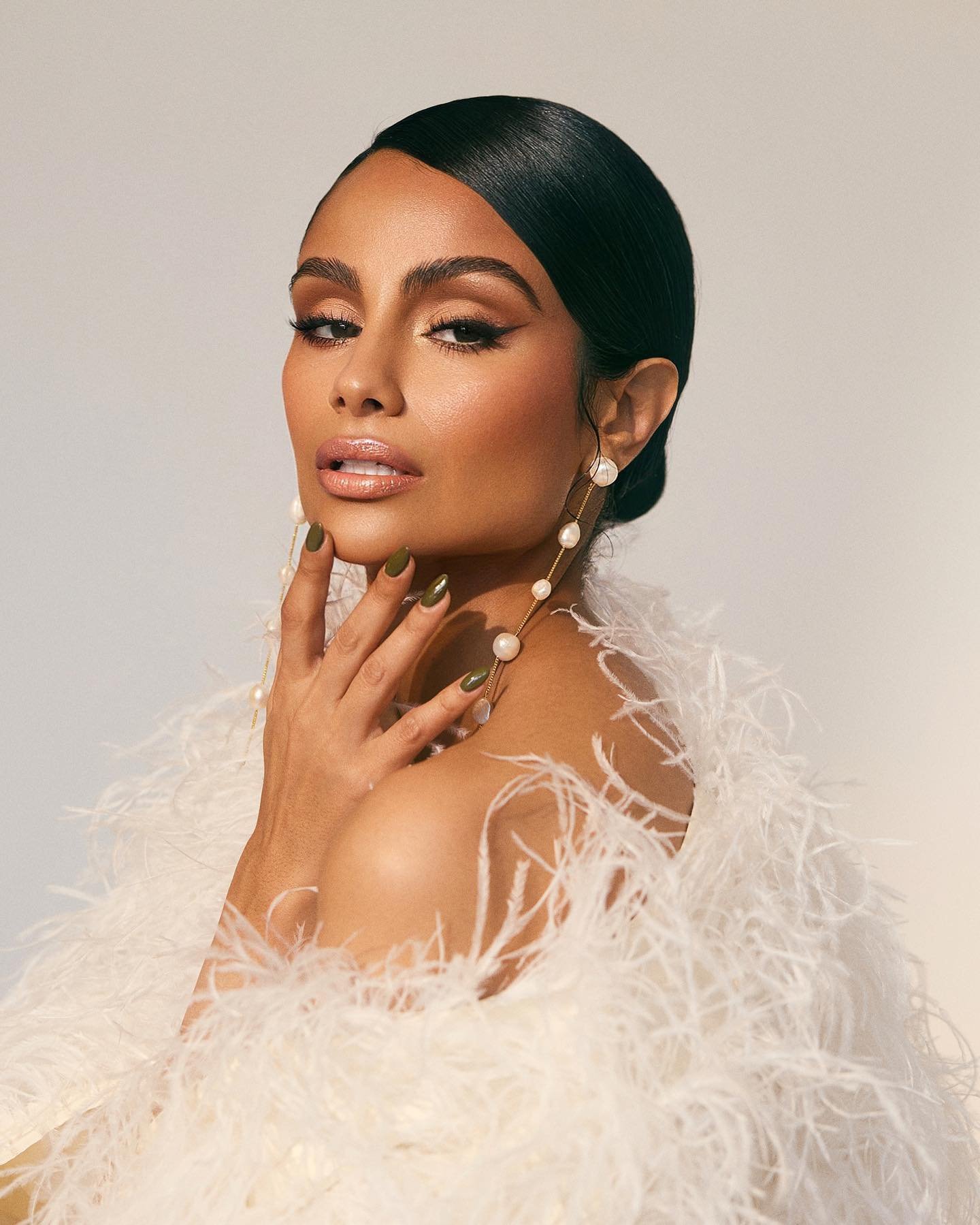 Close-up portrait of a brunette adult female model in a feathered white jacket and pearl earrings posing against a light gray backdrop.
