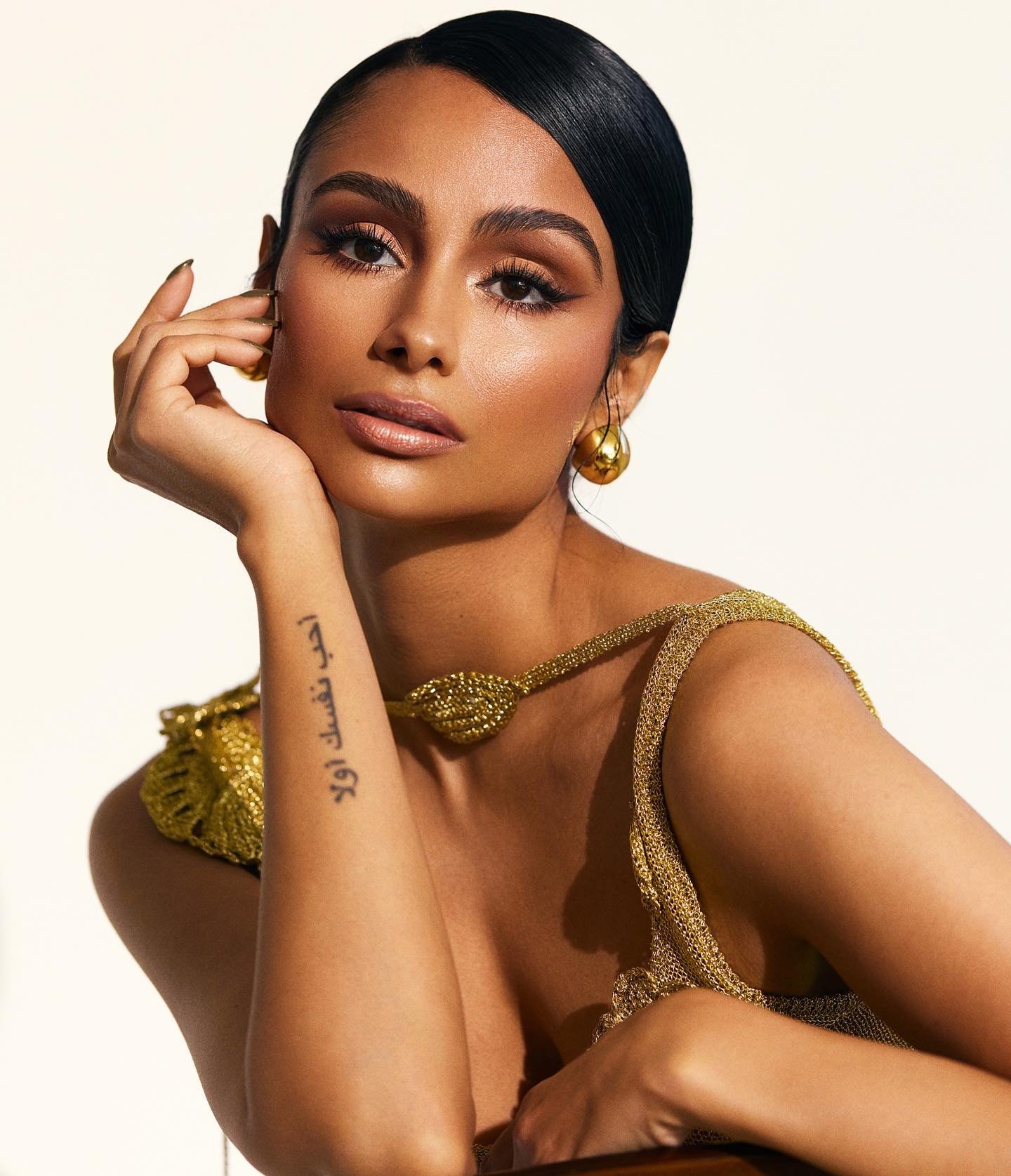 Close-up studio portrait of a light-skinned woman with sleek dark hair in an ornate gold dress and jewelry, gazing directly at the camera.