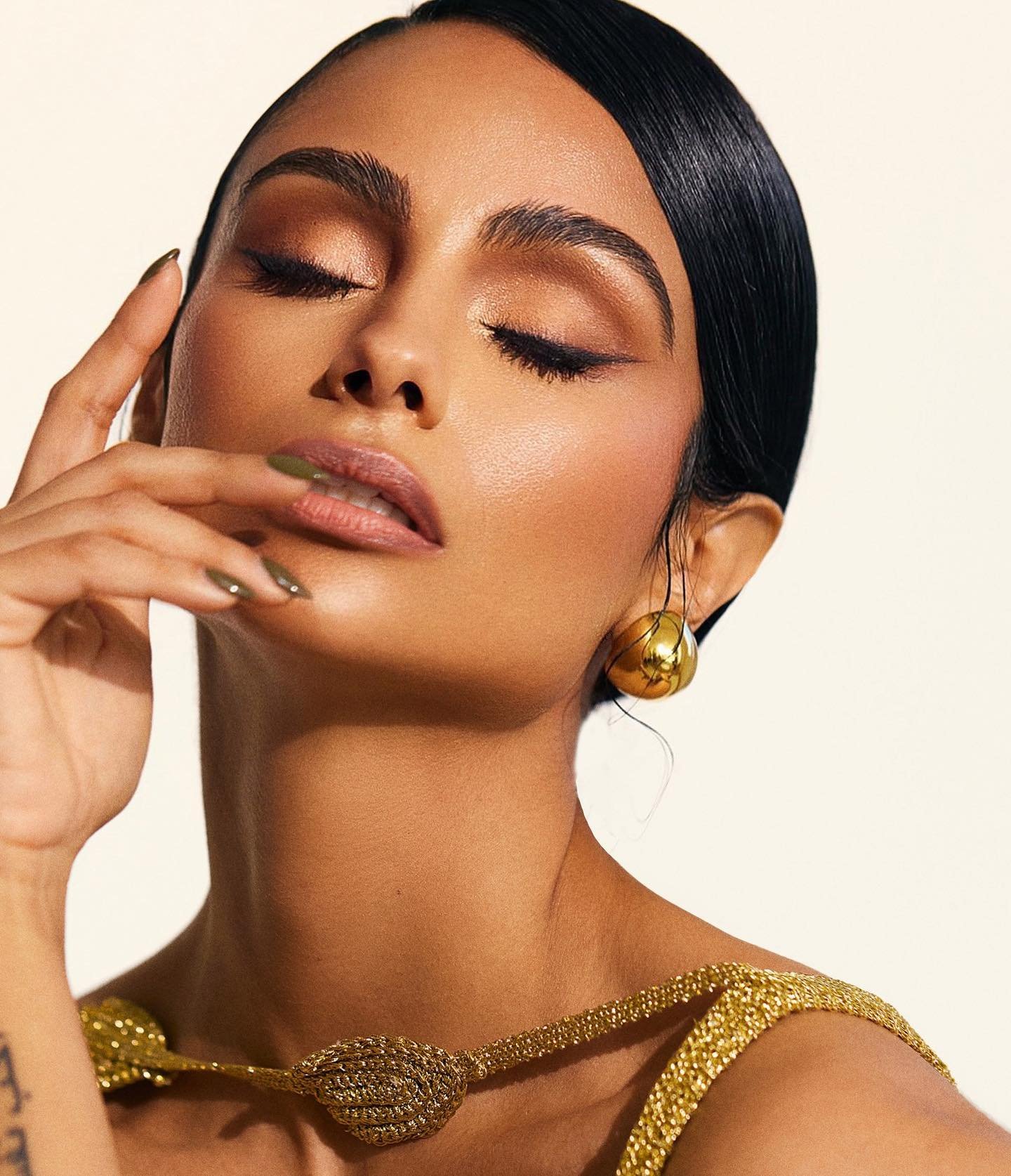 Close-up of a woman with brown skin, dark hair, and golden jewelry posing in front of a neutral background in a beauty style shot.