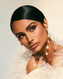 Close-up portrait of a woman with slicked-back dark hair wearing pearl drop earrings and a feather-trimmed garment.