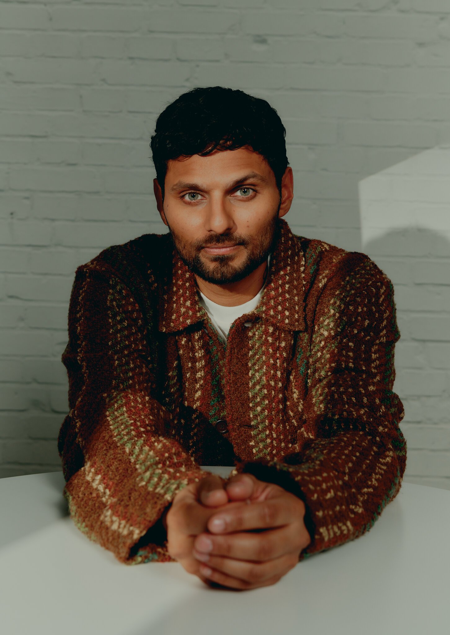 Portrait of a brown-haired man wearing a patterned jacket indoors with a neutral expression, set against a white brick wall and table.