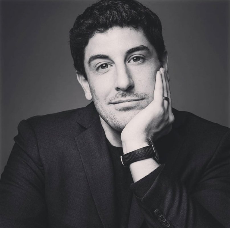 Monochrome portrait of a man with dark hair and a dark suit, resting his face on his hand in a contemplative pose against a grey background.