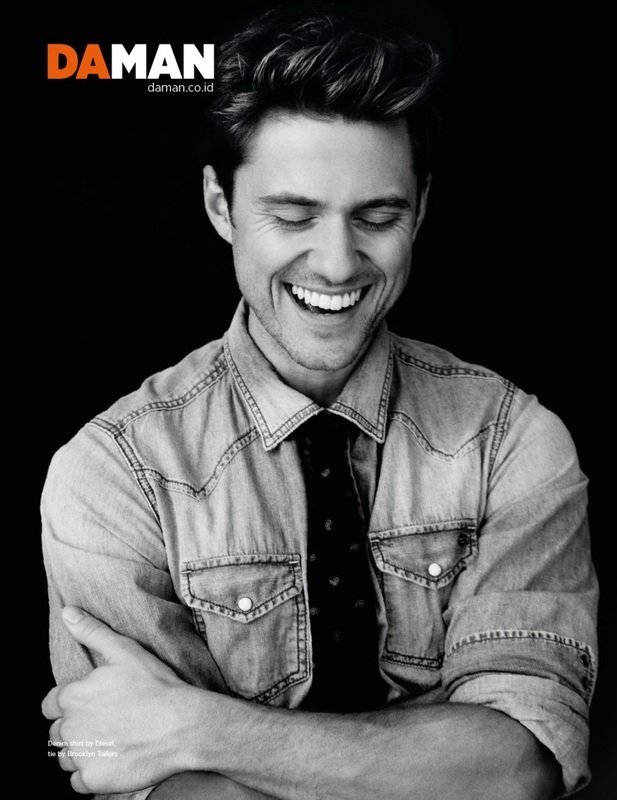 Black and white portrait of a smiling man with short hair wearing a denim shirt and patterned tie against a dark background.