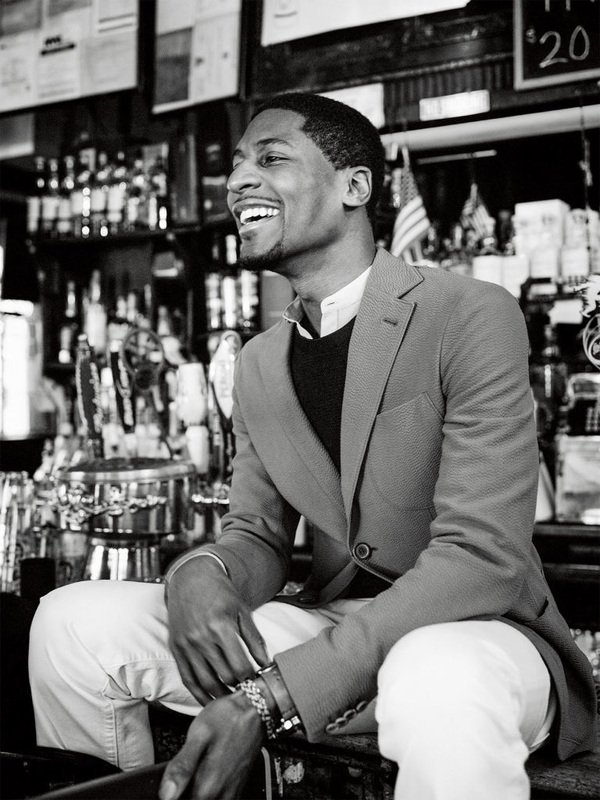 Black and white portrait of a smiling Black man in a blazer, seated at a bar with an array of bottles in the background.