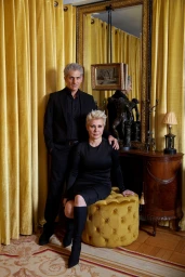 Man standing behind seated woman on tufted mustard stool, hair styled by Natasha Leibel in living room with yellow drapes.