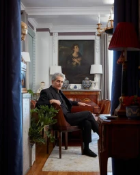 An older man seated in a refined living room, framed by dark blue curtains, with classic art and furnishings.