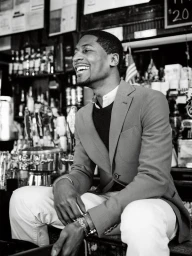 Black man smiling while seated in a bar interior with shelves of bottles in the background.