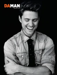 Black and white portrait of a smiling man with textured hair wearing a denim shirt against a dark background
