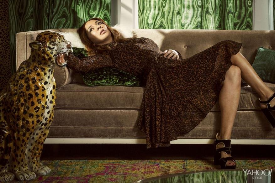 Woman in leopard print dress lounges on couch next to jaguar statue in a brightly lit room with patterned wallpaper and stylish decor.