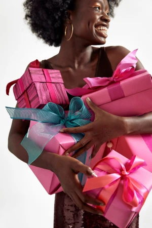 Smiling African American woman holding pink wrapped gifts with ribbons and bows. She wears a brown tank top against a white backdrop.