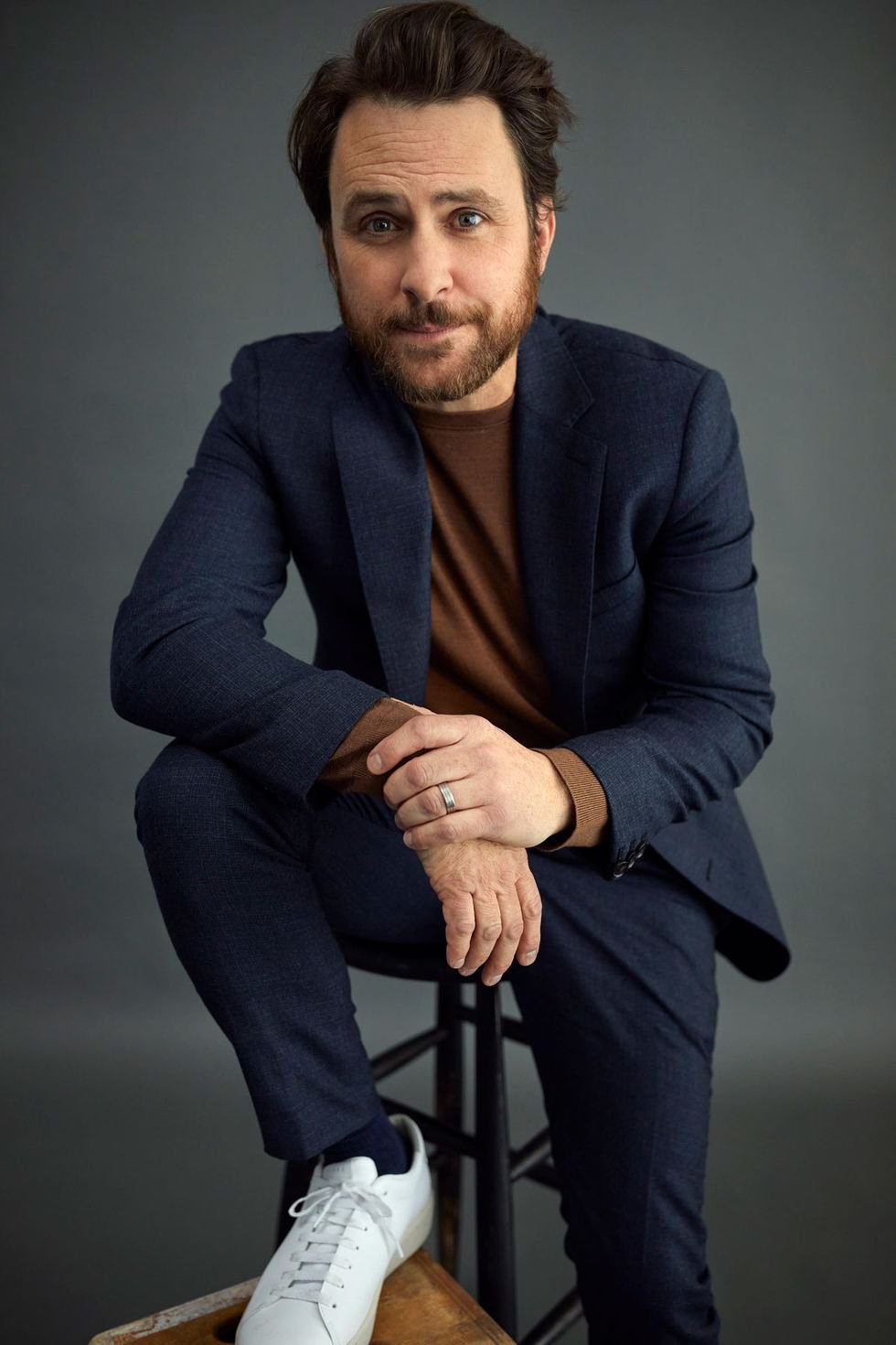 Portrait of adult male in a navy suit sitting on stool looking at the camera against a grey backdrop in a studio environment.