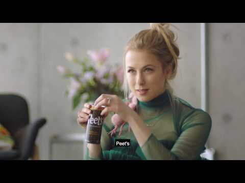Blonde woman in green top holds a can of Peet's coffee drink indoors, with flowers in the background, in a medium shot.