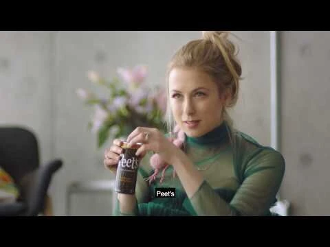 Blonde woman in green top holds a can of Peet's coffee drink indoors, with flowers in the background, in a medium shot.