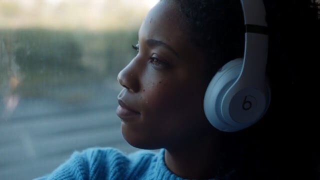 Close-up of a Black woman wearing white headphones, looking out the window on a moving train with a blurred landscape visible.