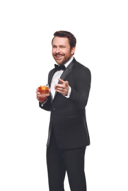 Adult male in a tuxedo smiles, points forward, and holds a cocktail glass on a white background in a medium shot. Beverage drink photography.