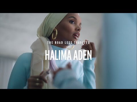 Halima Aden applies lip gloss in a close-up shot, wearing a headscarf, gold hoop earrings, and a light blue sweater indoors.