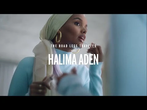 Halima Aden applies lip gloss in a close-up shot, wearing a headscarf, gold hoop earrings, and a light blue sweater indoors.